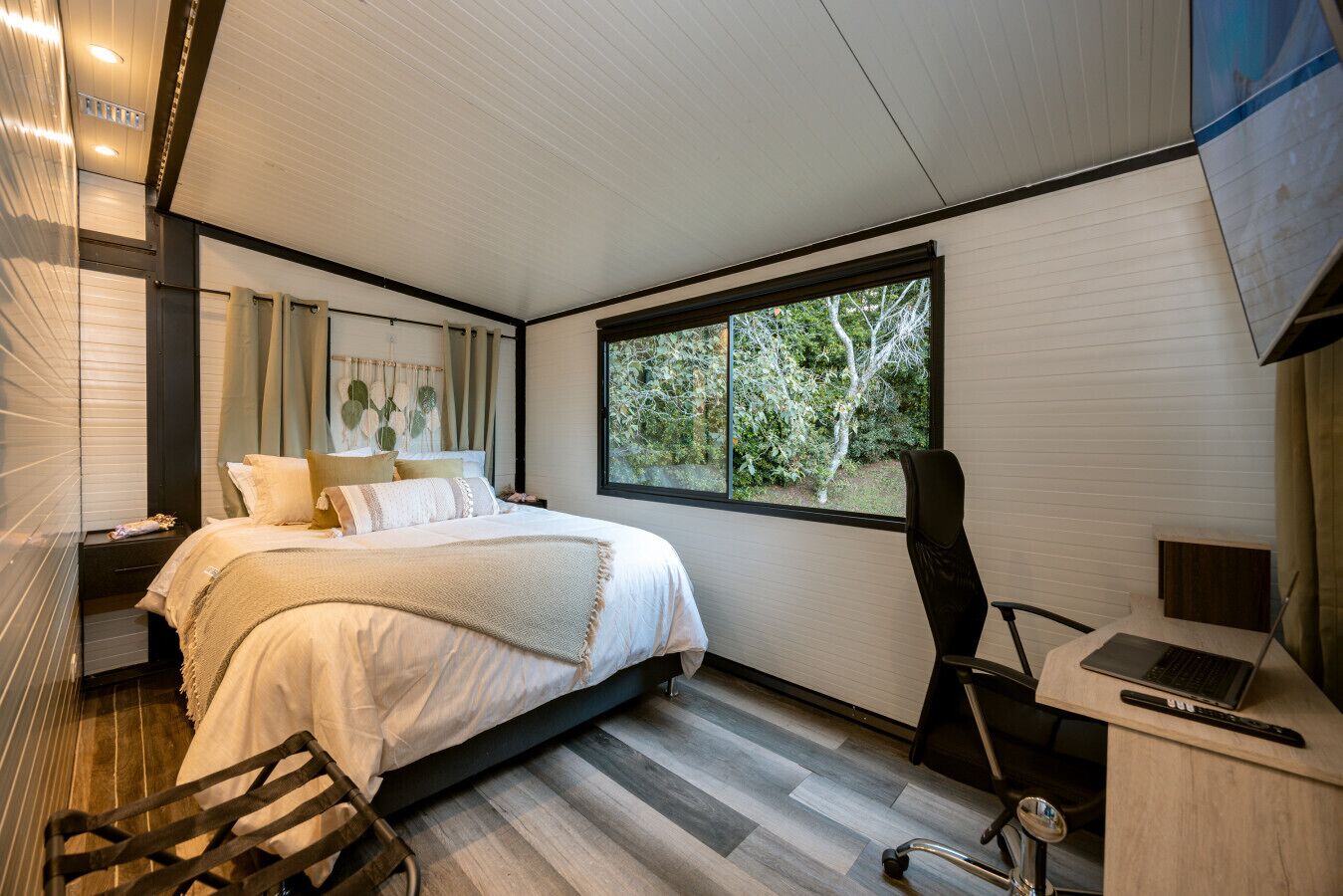 Un dormitorio moderno con una cama grande, un escritorio con una computadora portátil y un ventanal con vista a árboles.