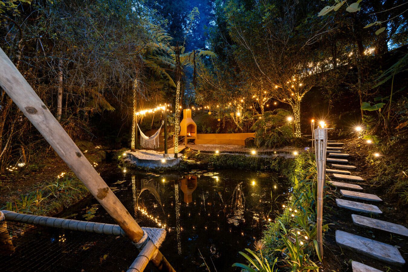 A serene outdoor setting with a wooden bridge over a reflective pond, surrounded by lush greenery and string lights.