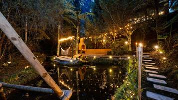 A serene outdoor setting with a wooden bridge over a reflective pond, surrounded by lush greenery and string lights.