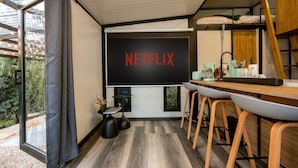 A modern room with a large TV screen displaying "NETFLIX," a wooden desk, and a dining area with bar stools.