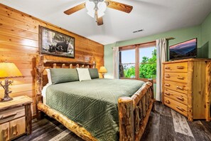 Quarto com decoração em madeira, com uma cama, cómoda de madeira, ventoinha no teto, janela com vista e um quadro emoldurado na parede.