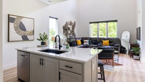 Een moderne keuken met een centraal eiland, een zithoek met een bank en stoelen, en een groot raam met uitzicht op groen.
