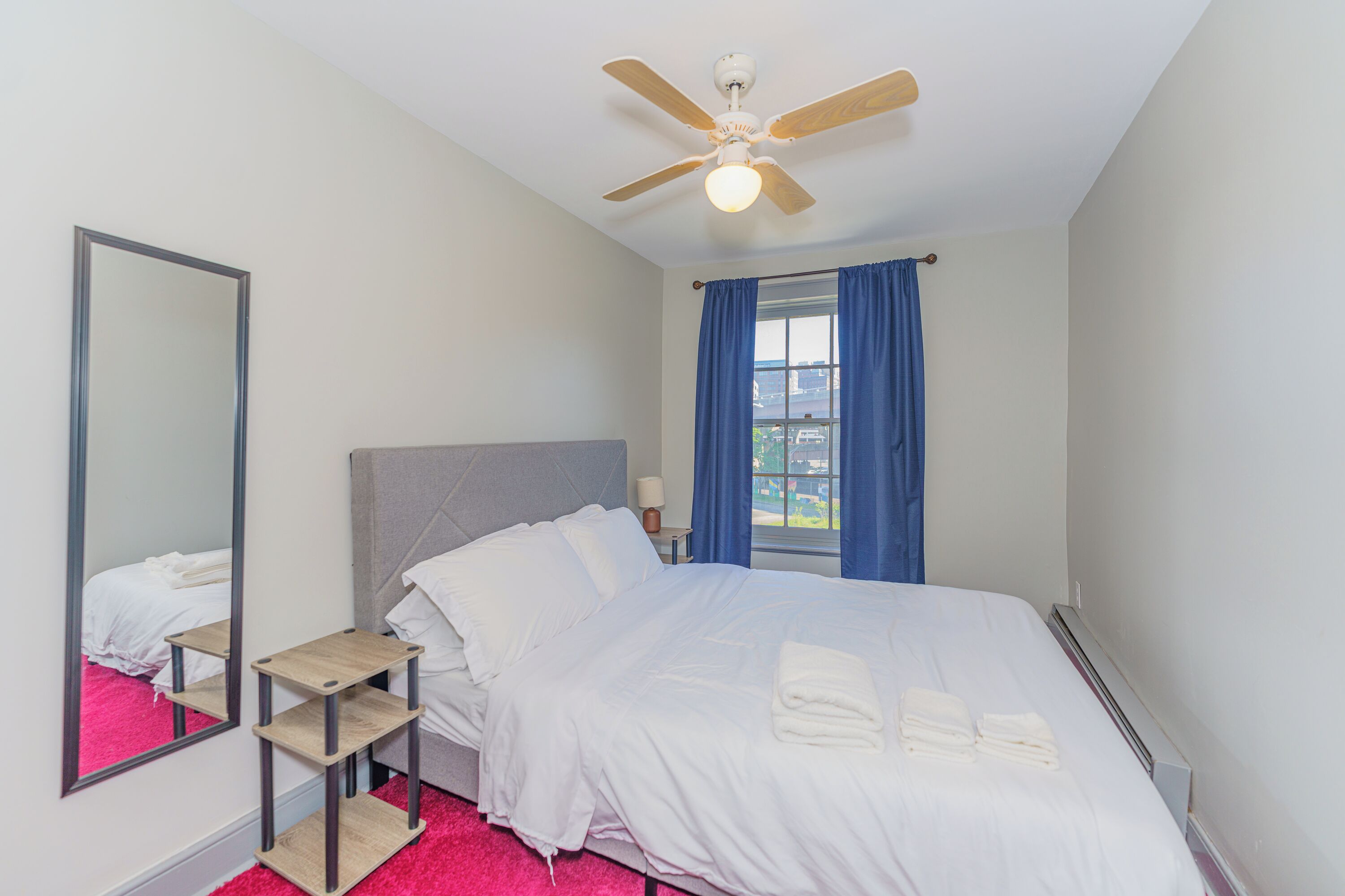 Ein Hotelzimmer mit Bett, Deckenventilator, Fenster mit blauen Vorhängen, Nachttisch und Spiegel.
