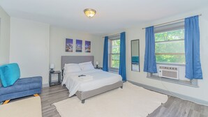A bedroom with a bed, a nightstand, a lamp, a window with curtains, a wall-mounted air conditioner, and a cityscape artwork on the wall. - Massive 5BD/4BA 3-Story Duplex @ Downtown Albany (Albany)