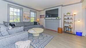 A modern living room with a grey sofa, a coffee table, and a TV. There is a wooden floor, a blue rug, and a bookshelf with plants. - Massive 5BD/4BA 3-Story Duplex @ Downtown Albany (Albany)