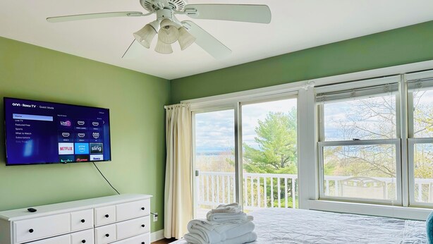 Une chambre avec un lit, une commode blanche, un ventilateur de plafond, un téléviseur affichant l’interface Roku TV et un balcon avec vue.