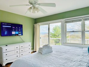 Une chambre avec un lit, une commode blanche, un ventilateur de plafond, un téléviseur affichant l’interface Roku TV et un balcon avec vue.