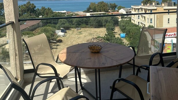 A balcony with a round table and chairs, overlooking a body of water and a hilly landscape. - Modern seaview apartment just 5 min from restaurants and markets, free parking!” (Vlora)