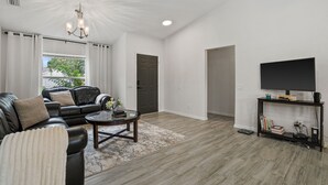 A modern living room with a leather sofa, a coffee table, a flat-screen TV on a stand, and a large window with curtains. - Serenity Heaven Gateway/Pool/Sauna/Golf course (Bradenton)