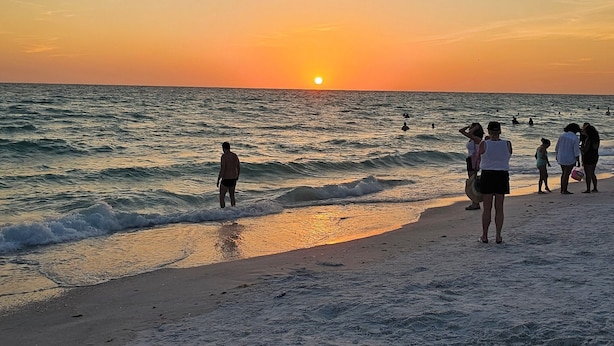 A beach scene with people walking along the shore and swimming in the water during sunset. - Serenity Heaven Gateway/Pool/Sauna/Golf course (Bradenton)
