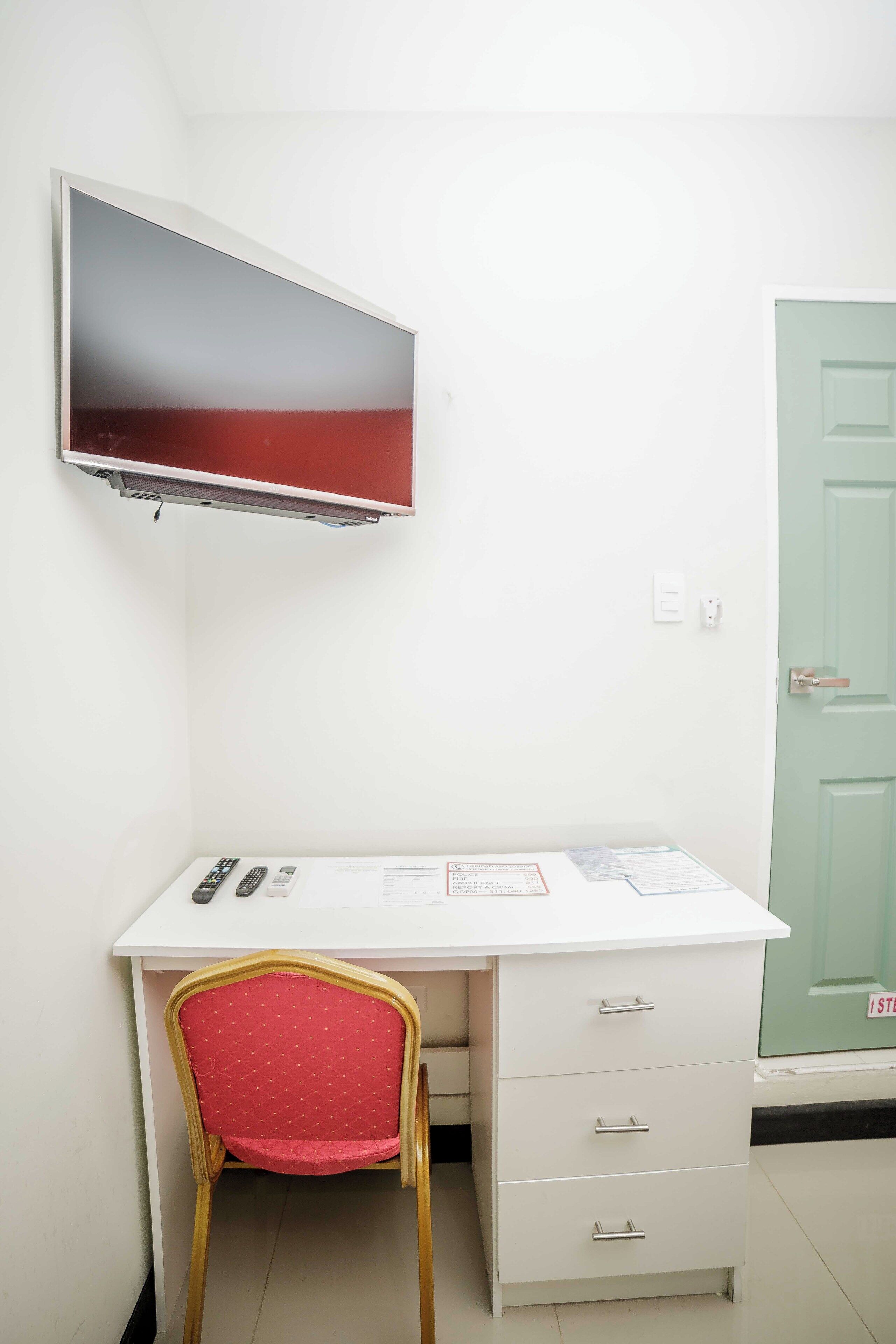 A white desk with drawers, a red chair, a flat-screen TV mounted above the desk, and a green door to the right.