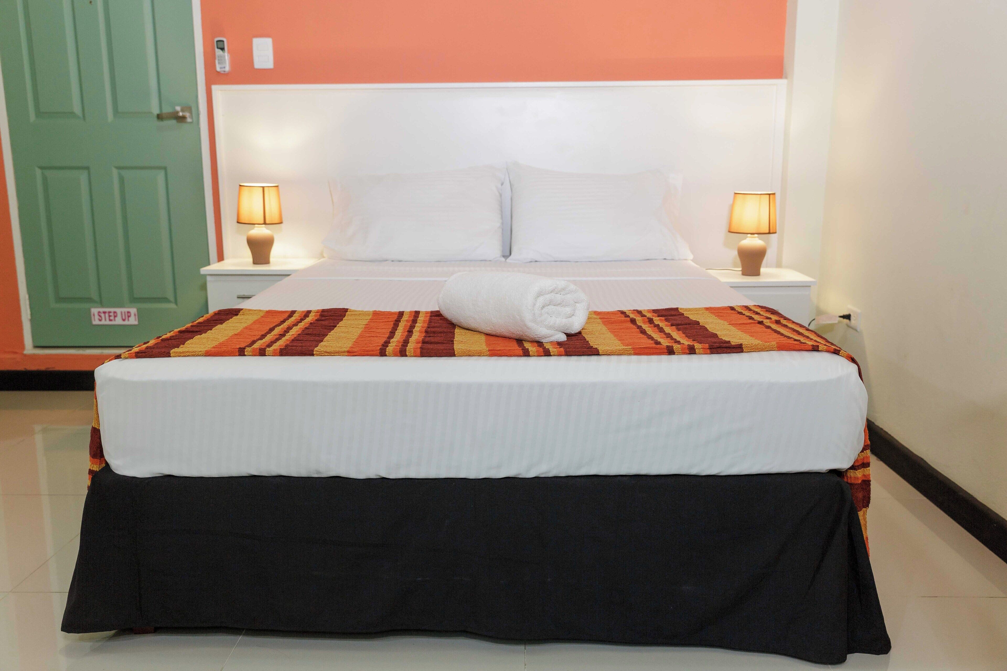 A neatly made bed with a white comforter and a rolled towel, two bedside lamps, and a green door with a 'Step Up' sign.