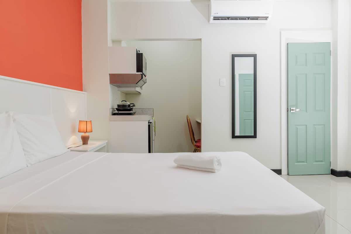 A hotel room with a bed, a nightstand with a lamp, a kitchenette, a door, and a mirror on the wall.