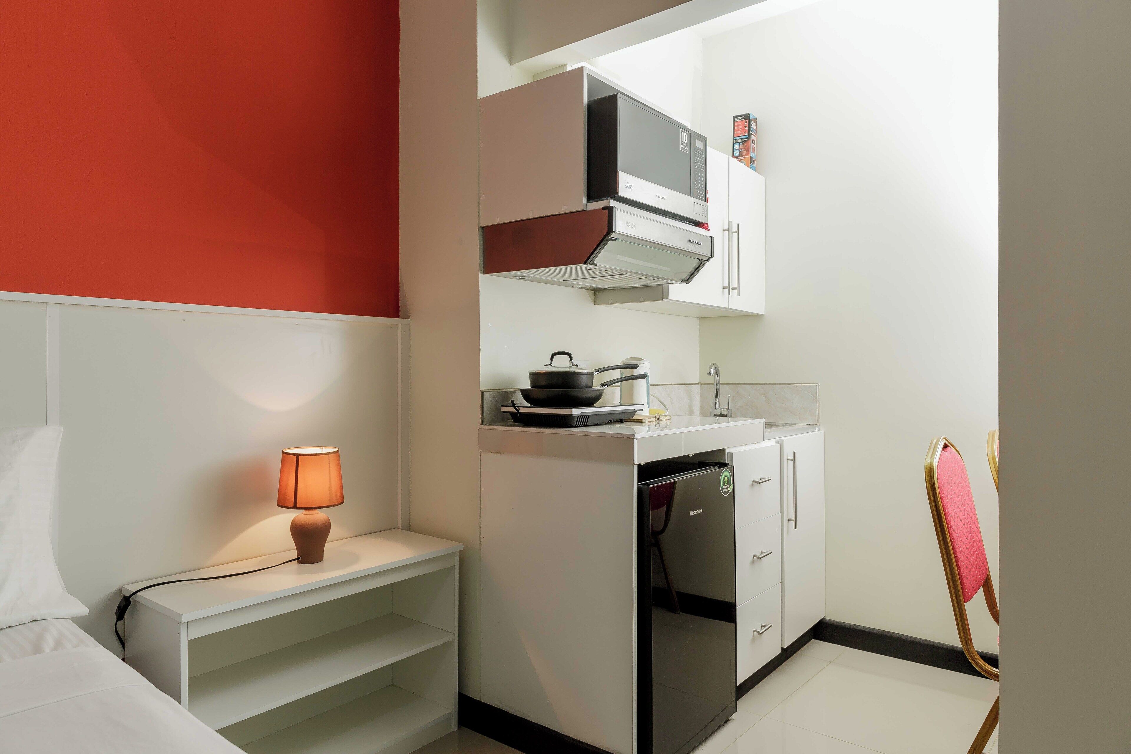 Une cuisine compacte équipée d’un four à micro-ondes, d’une cuisinière et d’un réfrigérateur. On y trouve une petite étagère avec une lampe, ainsi qu’une chaise dotée d’un coussin rose.