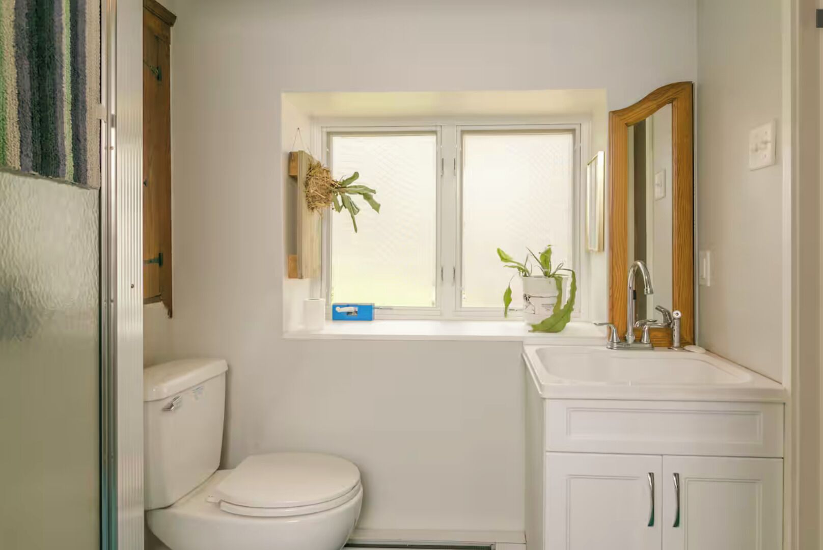 A small, white bathroom with a toilet, a window with a plant, and a sink with a mirror.