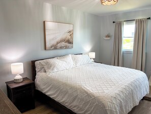 A bedroom with a large bed, two bedside lamps, a nightstand, a wall-mounted picture, and a window with curtains.