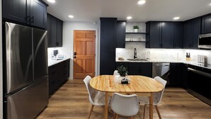 A modern kitchen with dark cabinets, a stainless steel refrigerator, a wooden dining table with white chairs, and a built-in microwave.