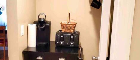 A kitchenette with a coffee maker, toaster, and basket on a cart, and a sign above that says "COFFEE, ANYONE?".
