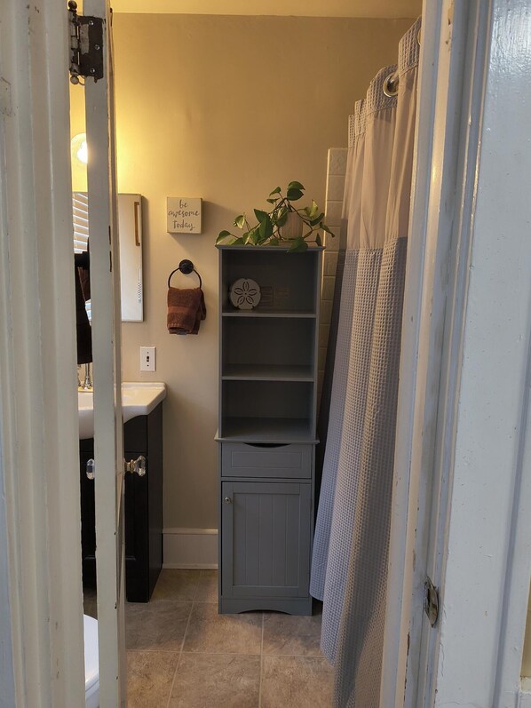 A bathroom with a white sink, a towel rack with a brown towel, a small plant on a shelf, and a shower curtain. - Robins Nest (Saranac Lake)