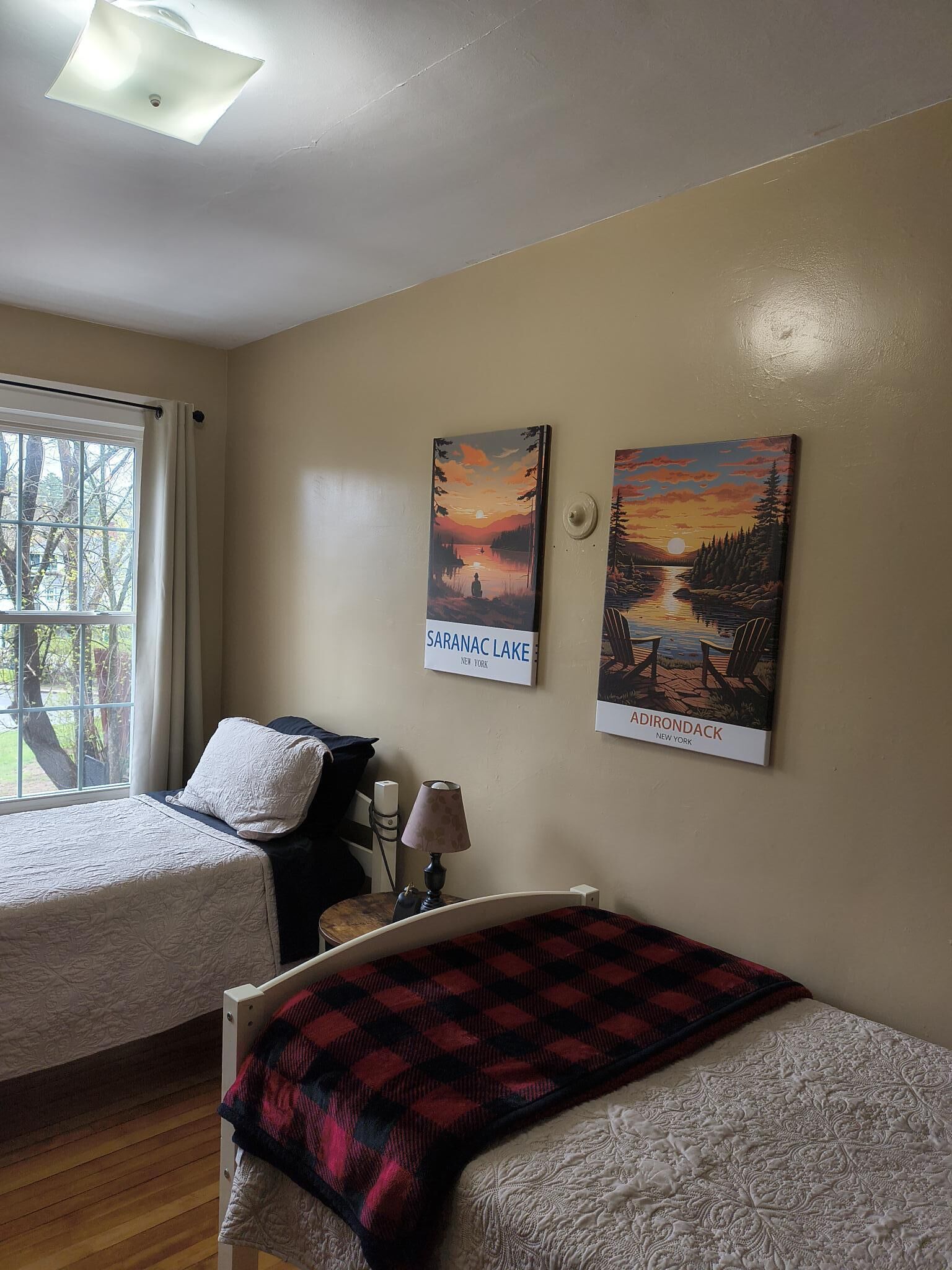 A room with two beds, a window with curtains, a lamp, and two paintings of lakes.