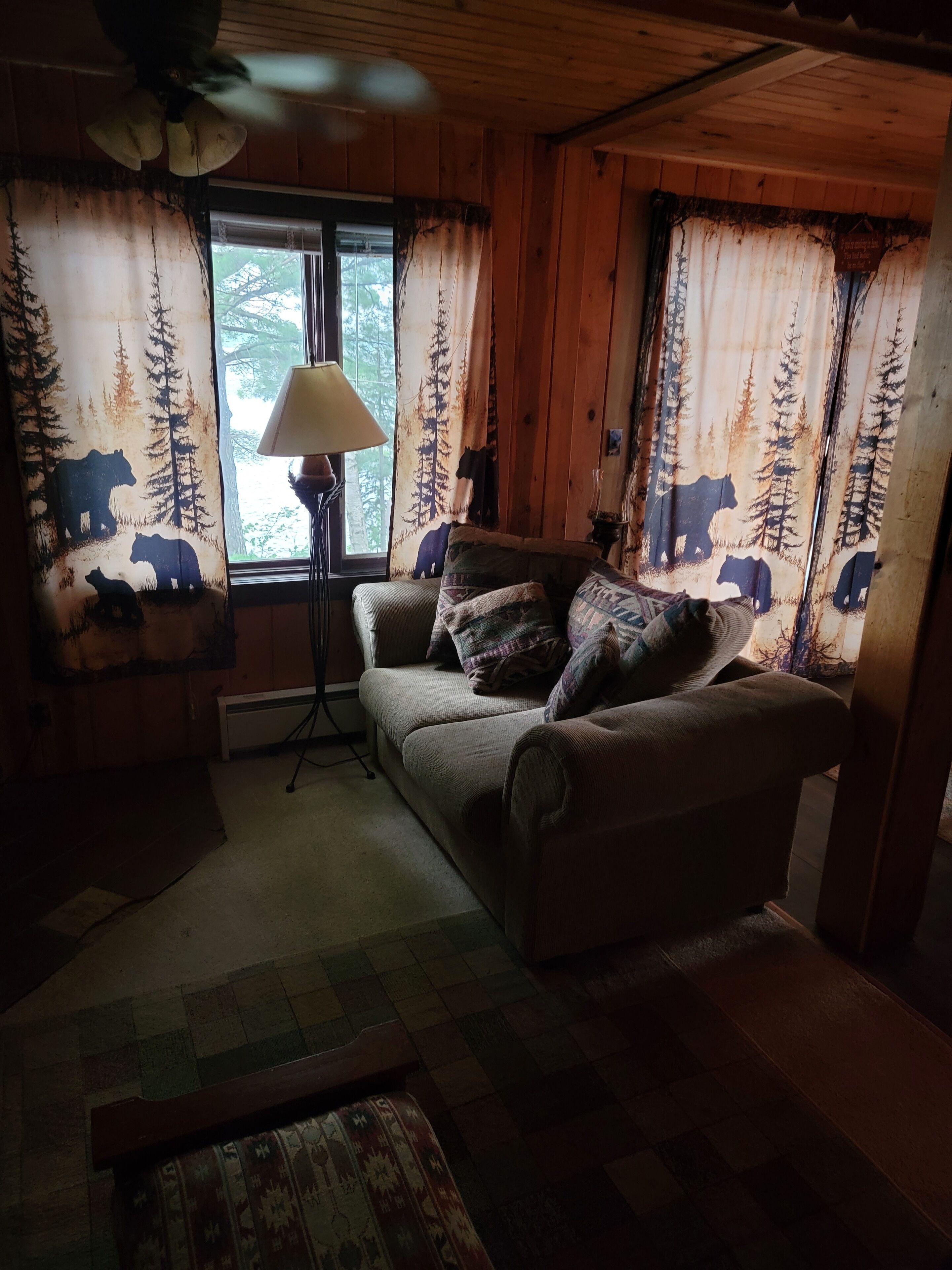 Interior acolhedor de cabana com teto de madeira, sofá com almofadas estampadas, luminária de chão e cortinas com desenho de urso e floresta.