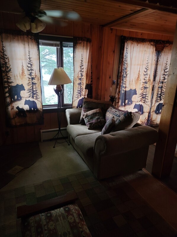 A cozy cabin interior with a wooden ceiling, a sofa with patterned cushions, a floor lamp, and curtains with a bear and forest design.