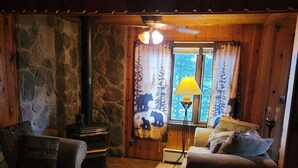 A cozy cabin interior with a stone wall, wooden furniture, a fireplace, and a view of trees through a window. - Charming 2-bedroom  lakefront cabin in delightful Phillip,WI (Phillips)
