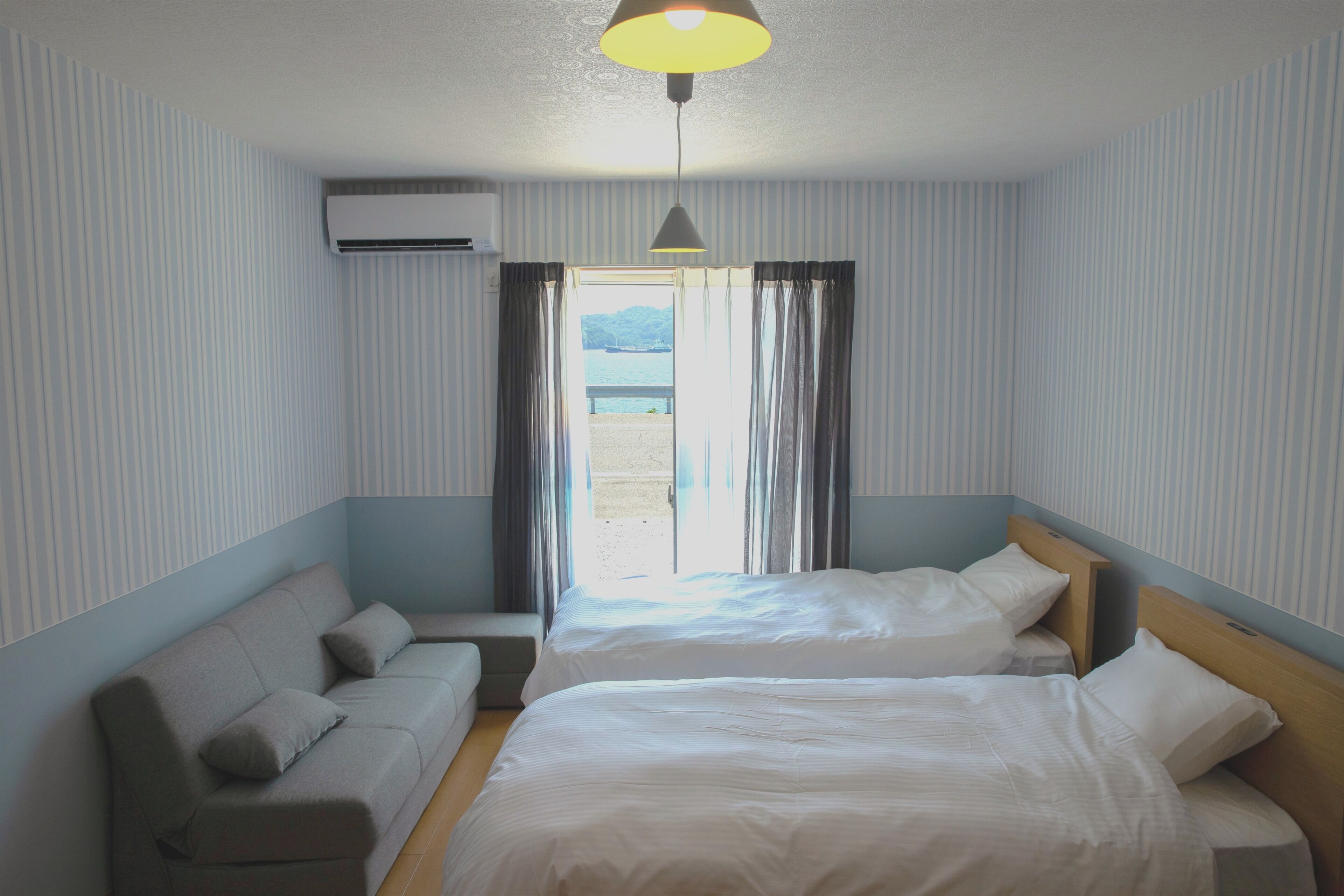 A hotel room with two beds, a sofa, an air conditioning unit, and a view of the outdoors.