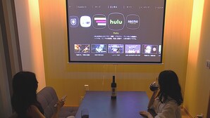 Two people seated at a table with a projector screen displaying streaming services. - SANTE 4073 STAY & SAUNA HAKATA ISLAND 1 (Imabari)