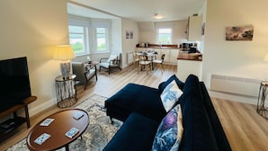 Een moderne woonkamer met een bank, televisie en salontafel. Er is een eethoek met stoelen en een tafel. De keuken is op de achtergrond zichtbaar.