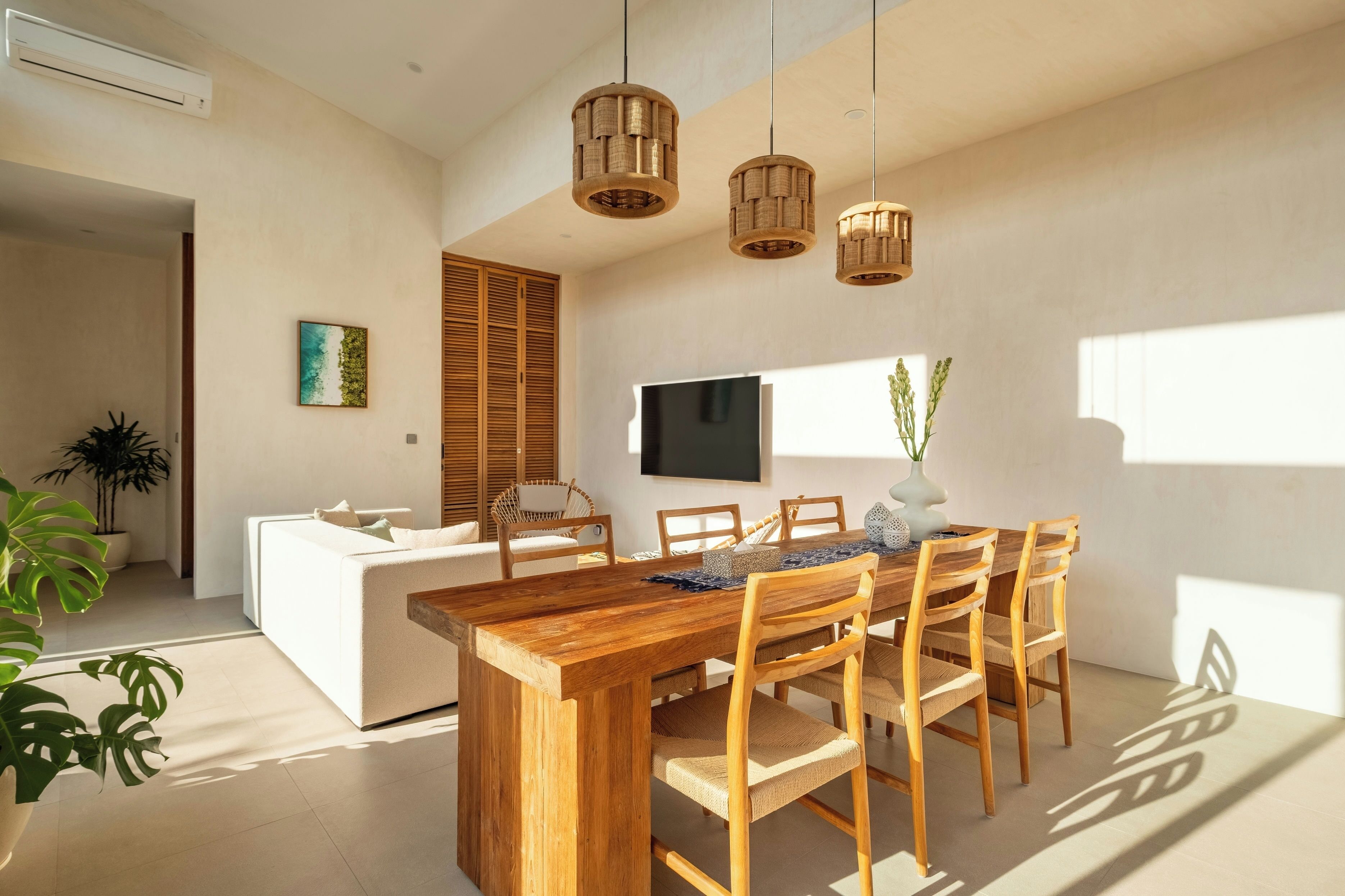 A modern living room with a wooden dining table, white sofa, and a flat-screen TV mounted on the wall.