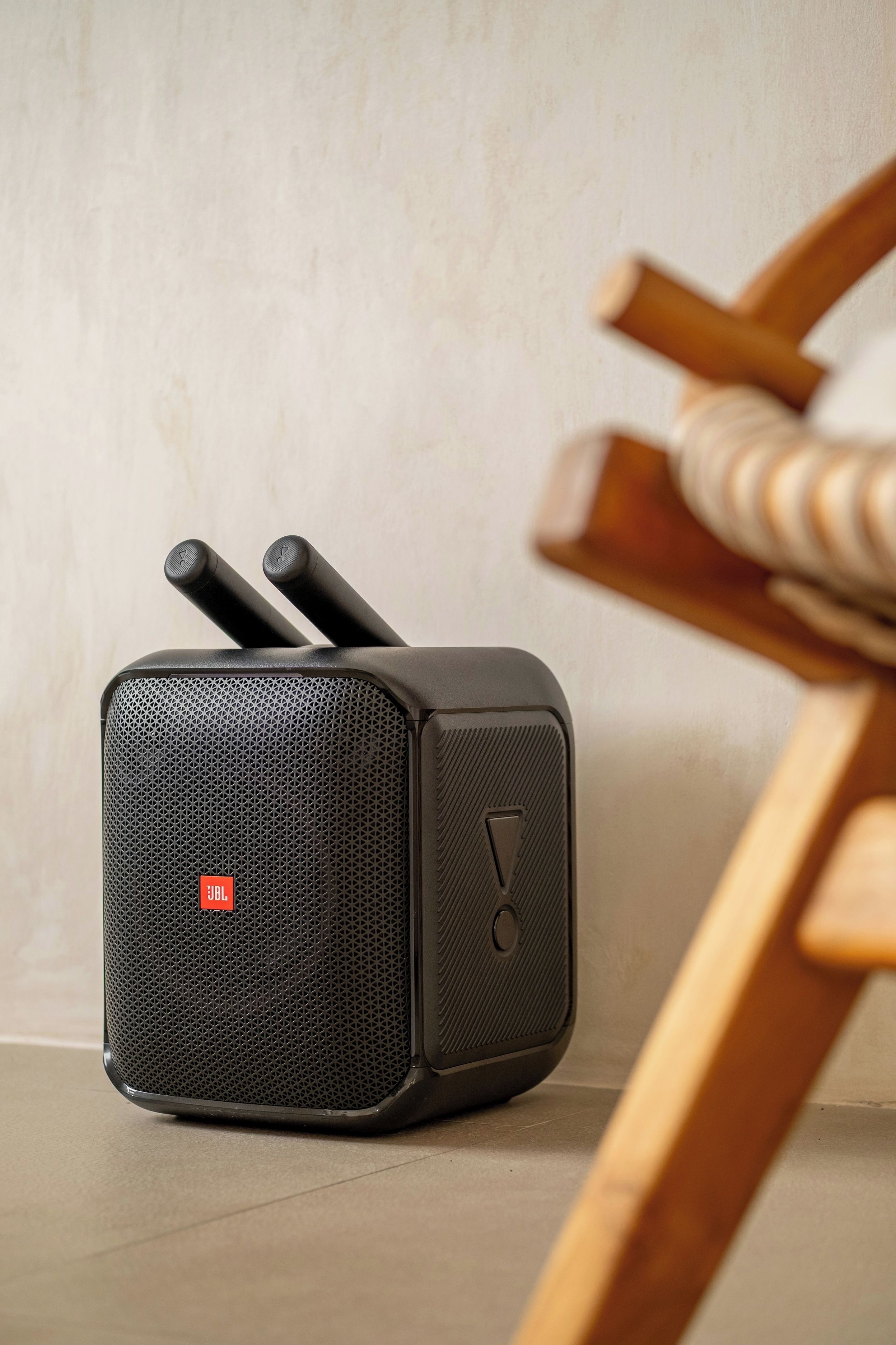 A black portable speaker with JBL branding, placed on a tiled floor next to a wooden chair.