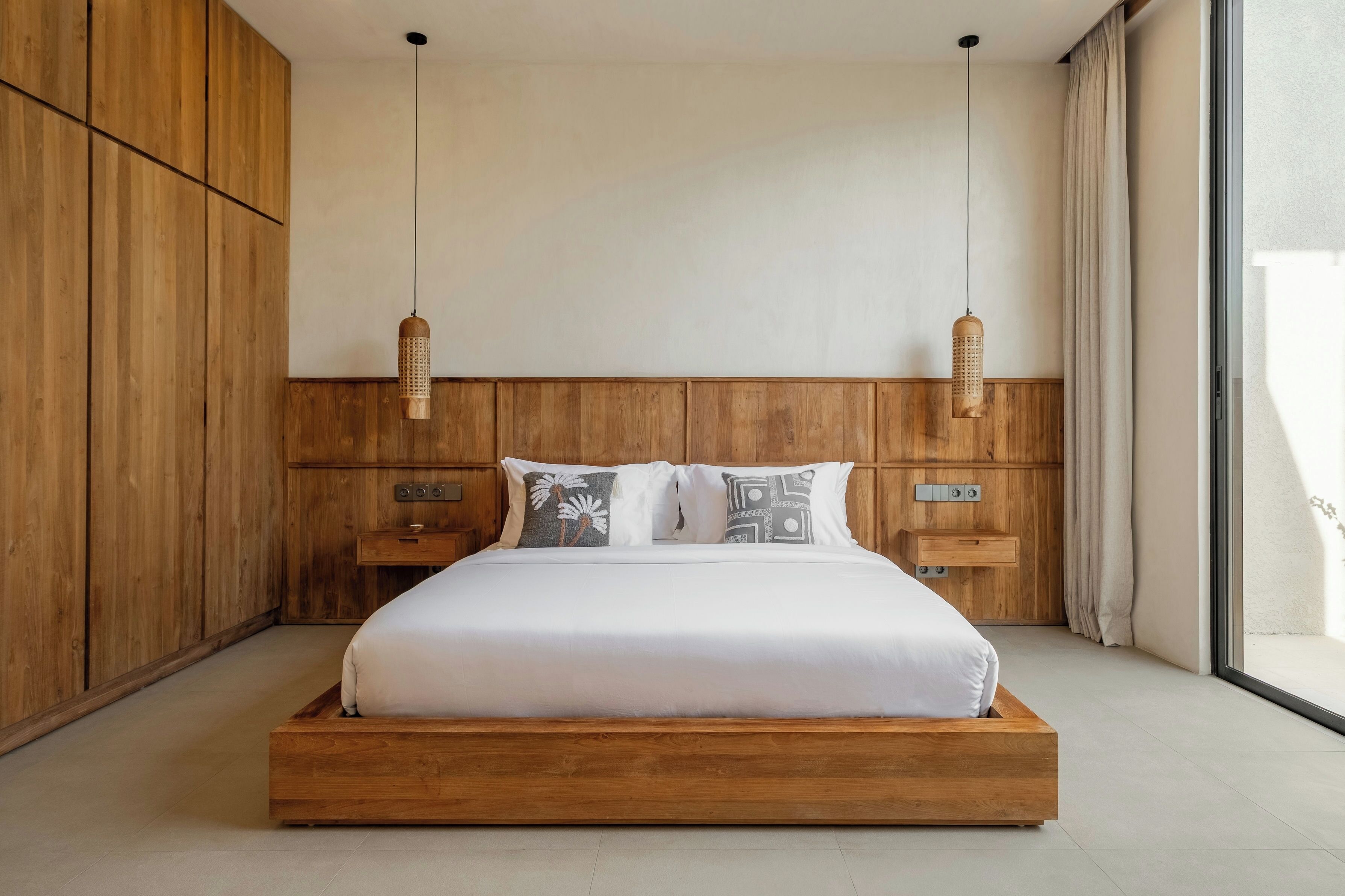A modern bedroom with a wooden headboard, a large bed with white bedding, two bedside tables, and a large window with curtains.