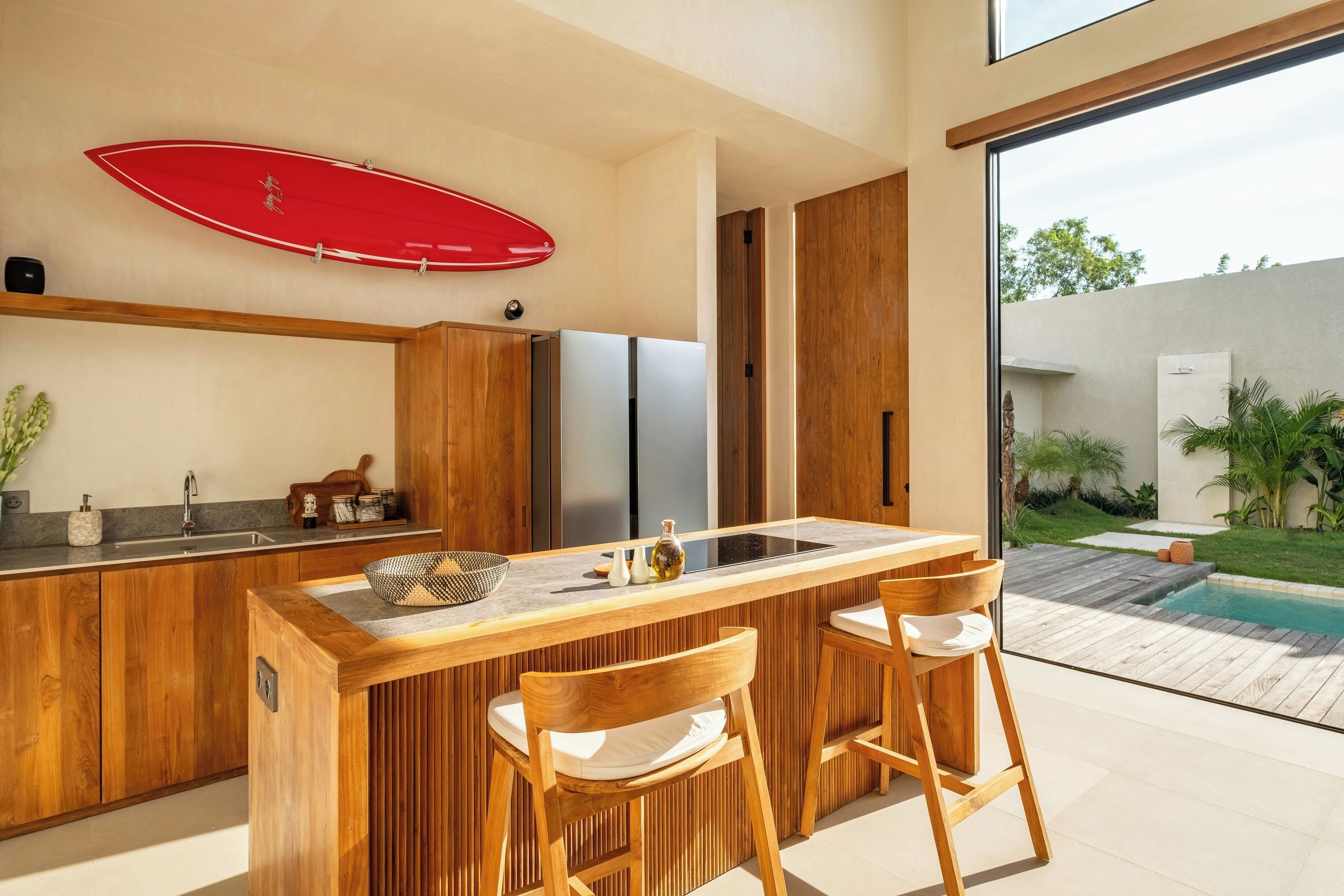 Eine moderne Küche mit Holdekor, einer Mittelinsel mit Barhocker, einem roten Surfbrett an der Wand und Blick auf einen Pool.