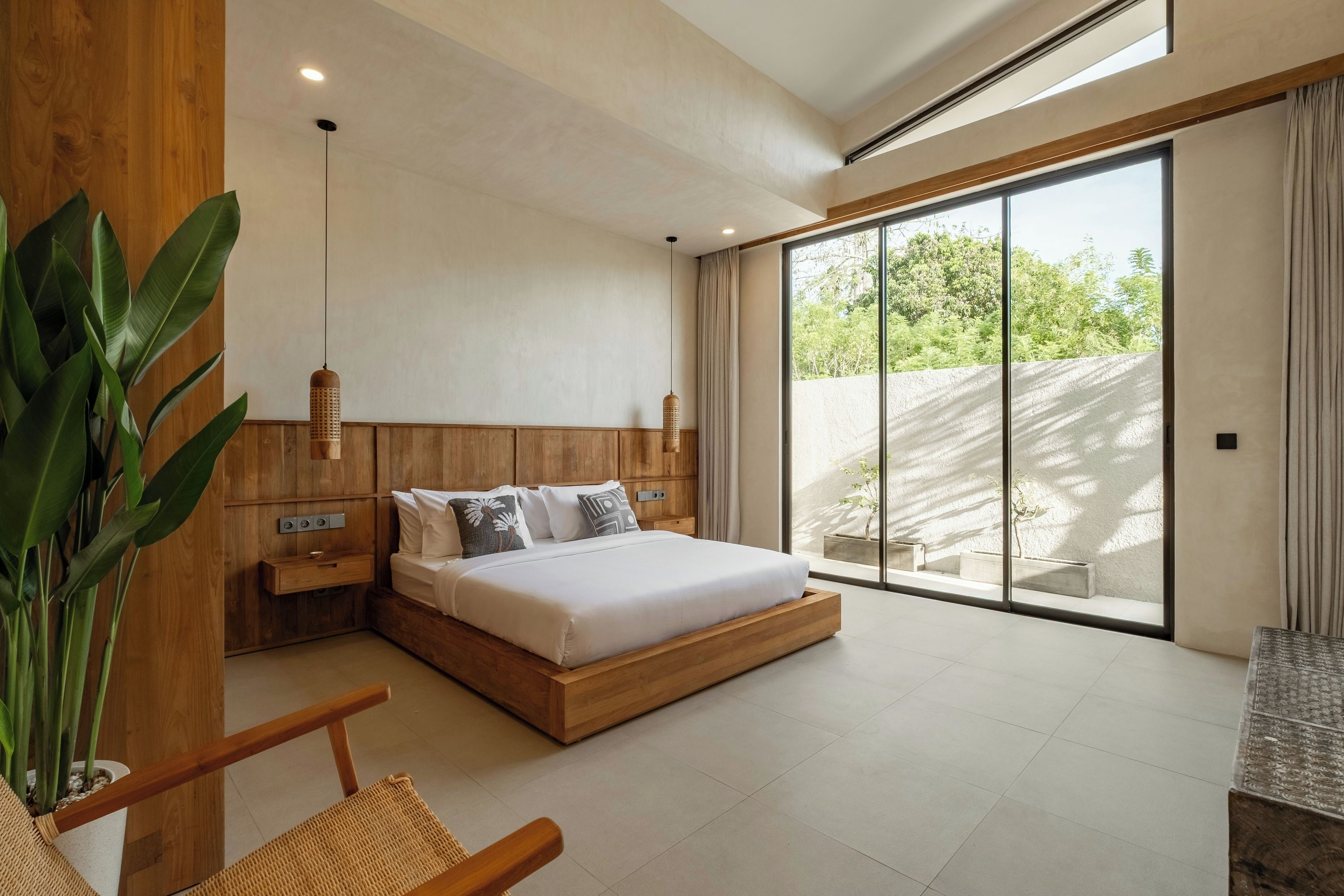 Un dormitorio moderno con cabecera de madera, cama grande, mesita de noche, lámpara colgante y vistas a la vegetación a través de amplias puertas correderas.
