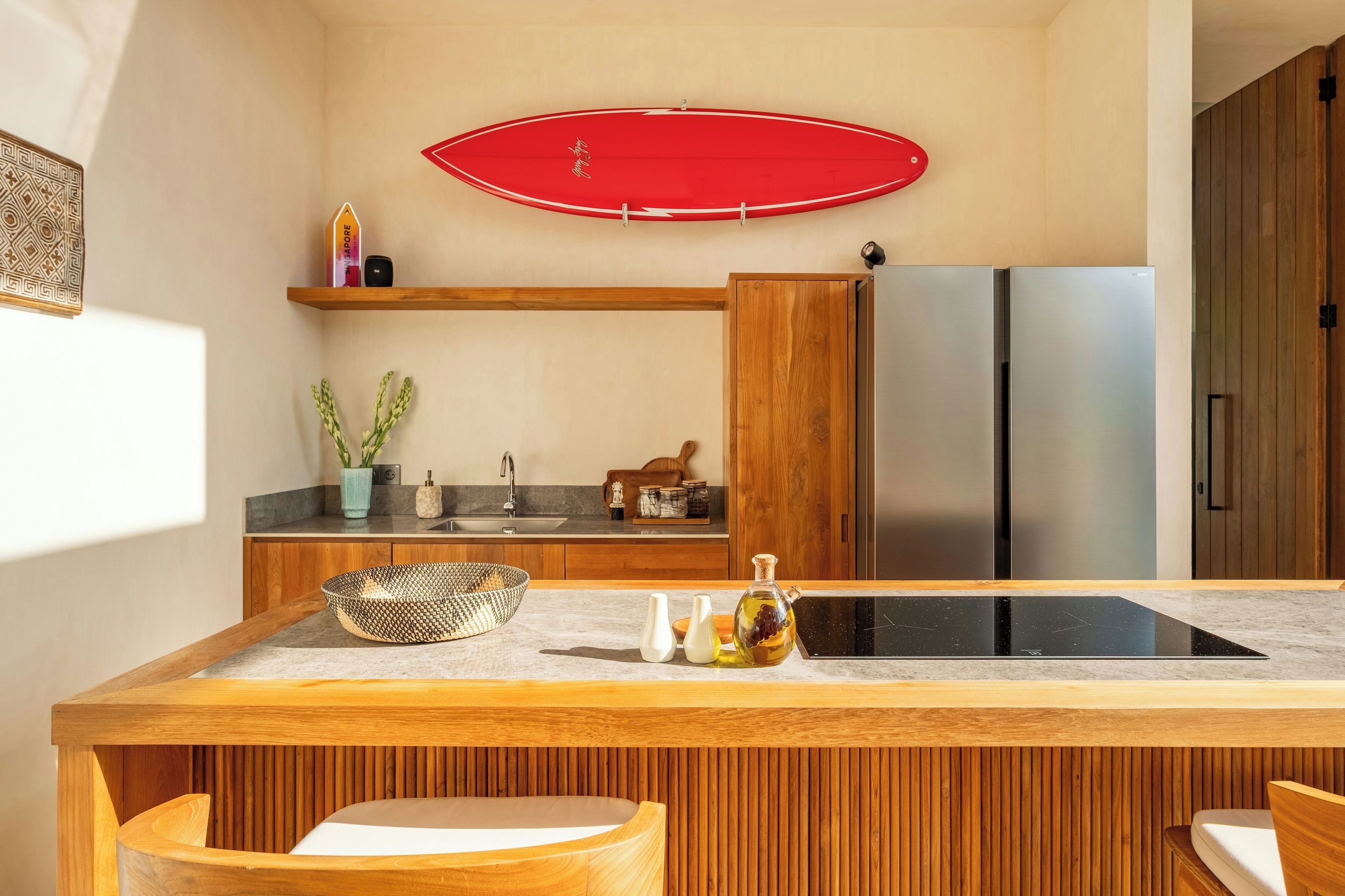 A modern kitchen with wooden cabinetry, a central island with a sink, and a red surfboard on the wall.