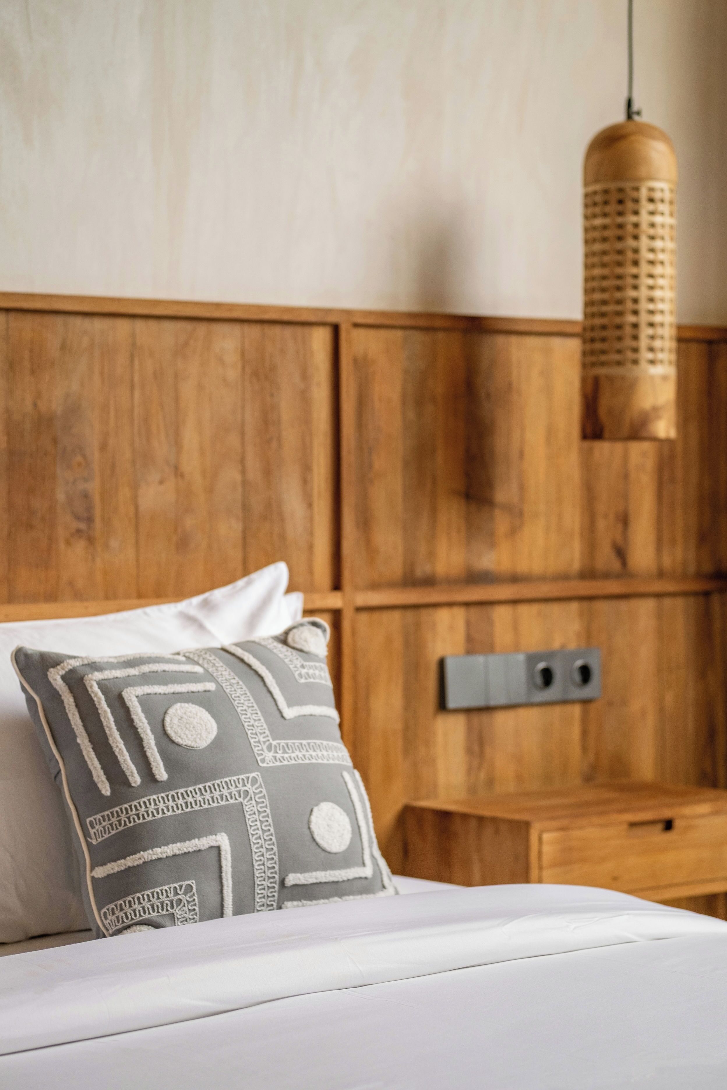 A bedroom with a wooden headboard, a bed with white bedding, a patterned pillow, and a hanging light fixture.