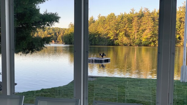 A lakeside view from a covered deck with a table and chairs, overlooking a calm lake and a dock with a small boat.