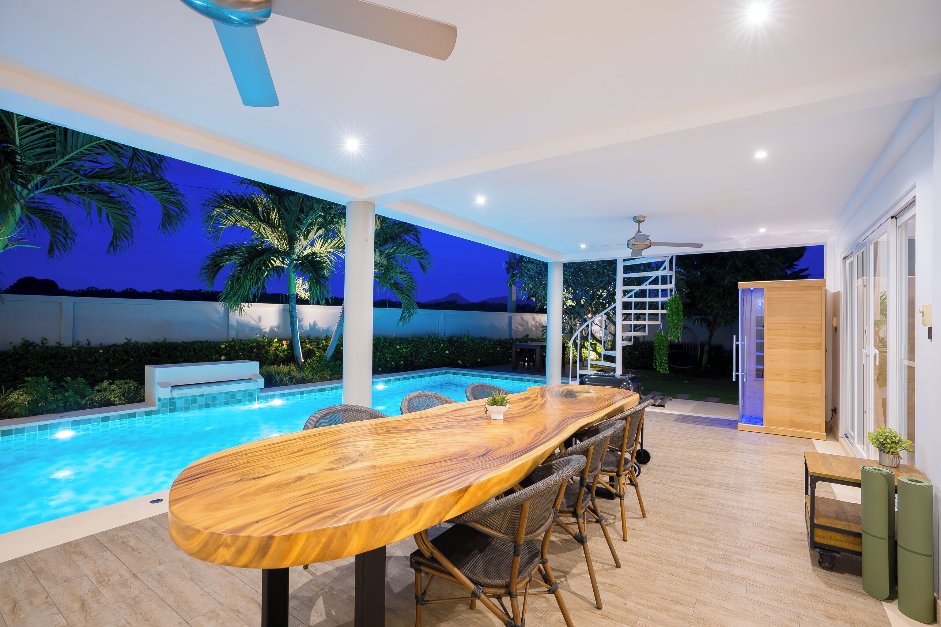 A modern outdoor pool area with a wooden table and chairs, surrounded by palm trees and a view of the mountains at dusk.