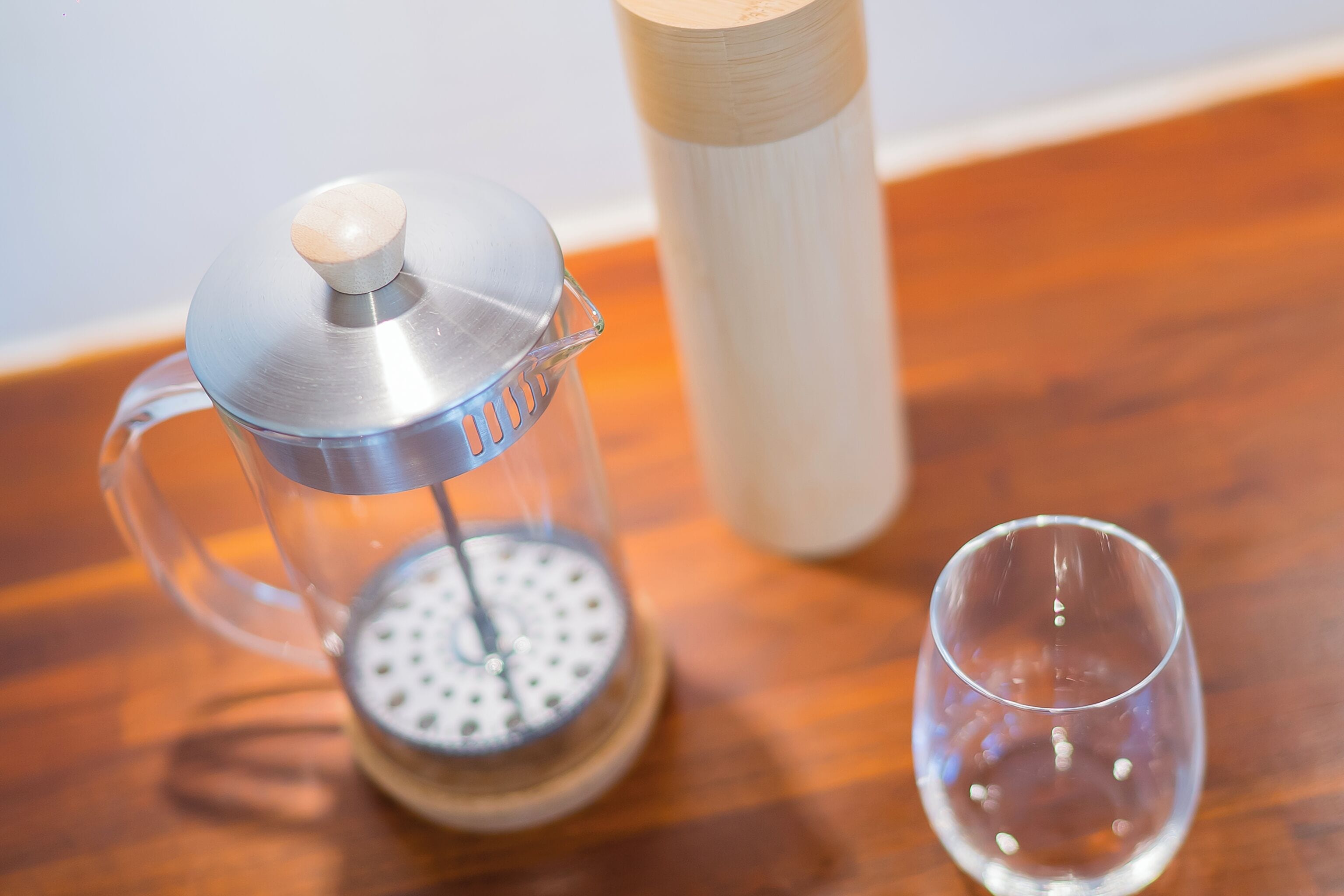 Una cafetera francesa, un vaso y un recipiente de madera sobre una mesa de madera.