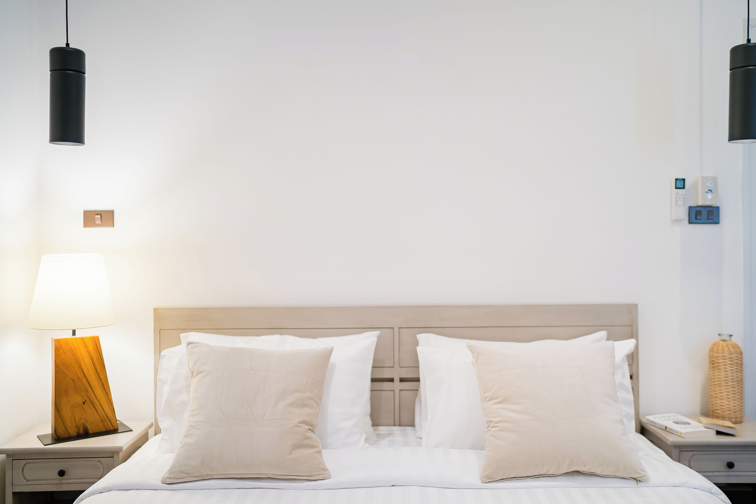 A neatly made bed with white linens and two beige pillows, a wooden bedside table with a lamp, and a modern pendant light.