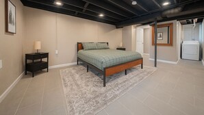 Un dormitorio con cama, mesitas de noche, alfombra y lavadora en una esquina.