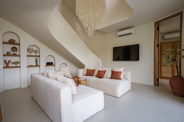 A modern living room with a white sofa set, a chandelier, and a flat-screen TV mounted on the wall.