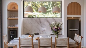 Una cocina moderna con encimera de mármol, una mesa de comedor con sillas de mimbre, horno empotrado y un amplio ventanal con luces colgantes.