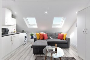 Een modern appartement met een grijze bank, een ronde glazen salontafel, een wasmachine en een dakraam.