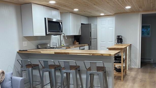 A modern kitchen with wooden ceiling, white cabinets, a central island with bar stools, and a grey sofa. - Tranquil cabin with WiFi in charming Au Train. (Au Train)