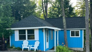 A small, single-story house with a blue exterior and a dark roof. There are two white chairs outside, a black trash bin, and a ladder leaning against the house. - Tranquil cabin with WiFi in charming Au Train. (Au Train)