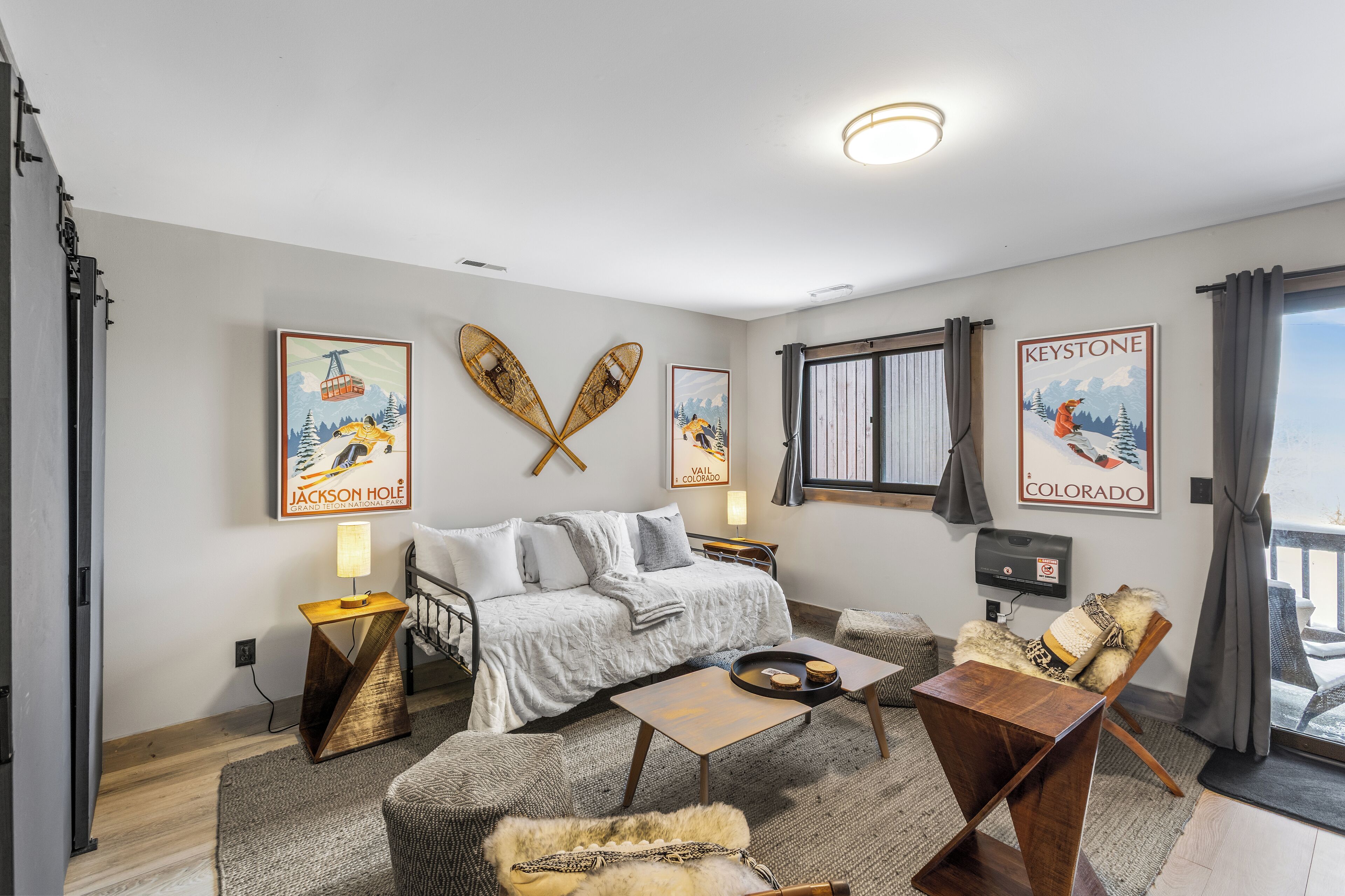Une chambre d’hôtel confortable avec un lit, une table de chevet en bois, un canapé et un fauteuil. Il y a deux téléviseurs fixés au mur et une fenêtre avec des rideaux.