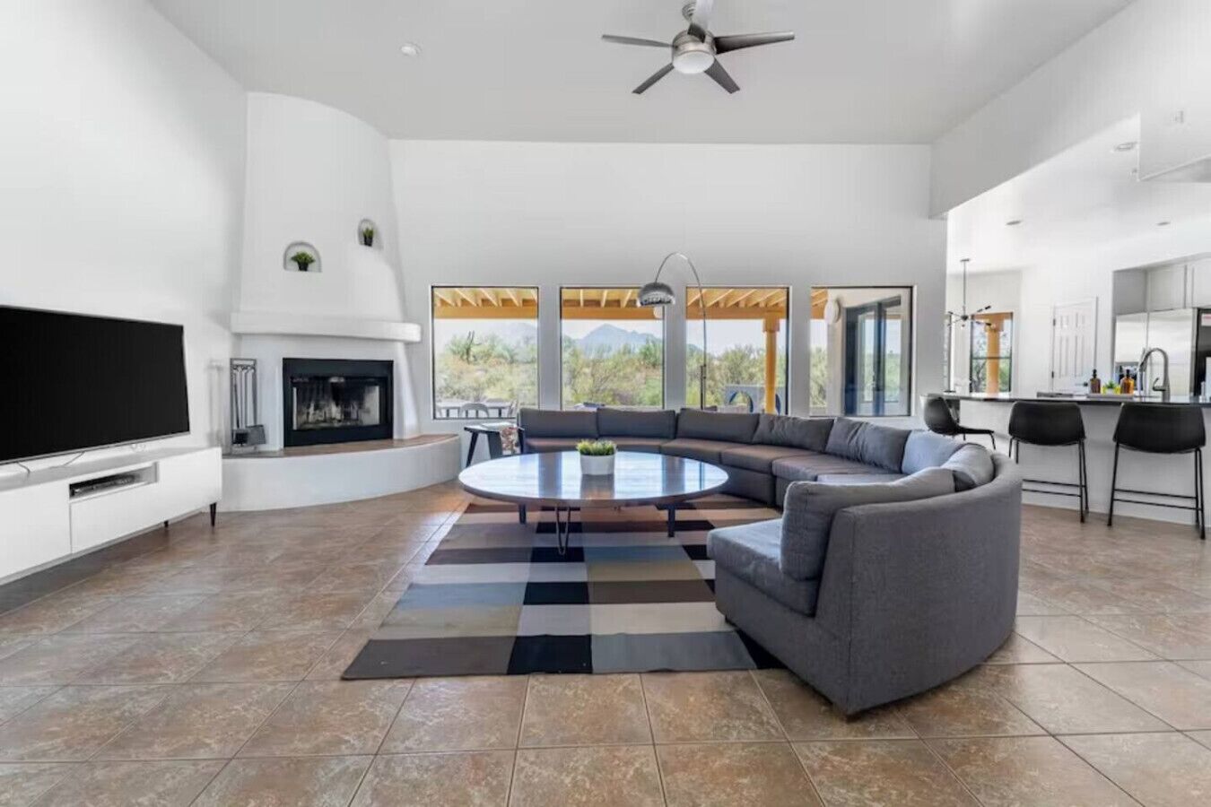 A spacious living room with a large flat-screen TV, a fireplace, a round coffee table, and a gray sectional sofa.