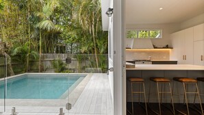 Een moderne keuken met een bar, een zwembad en weelderige tropische beplanting zichtbaar door een glazen deur.
